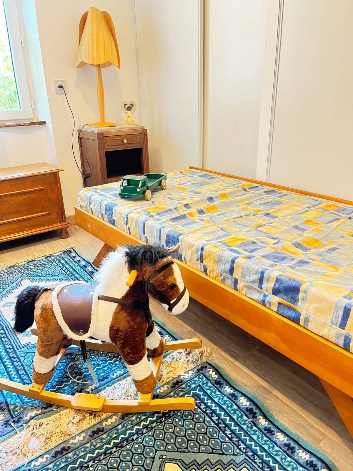 Een slaapkamer met een bed, een schommelpaard, een lamp en een klein houten nachtkastje.