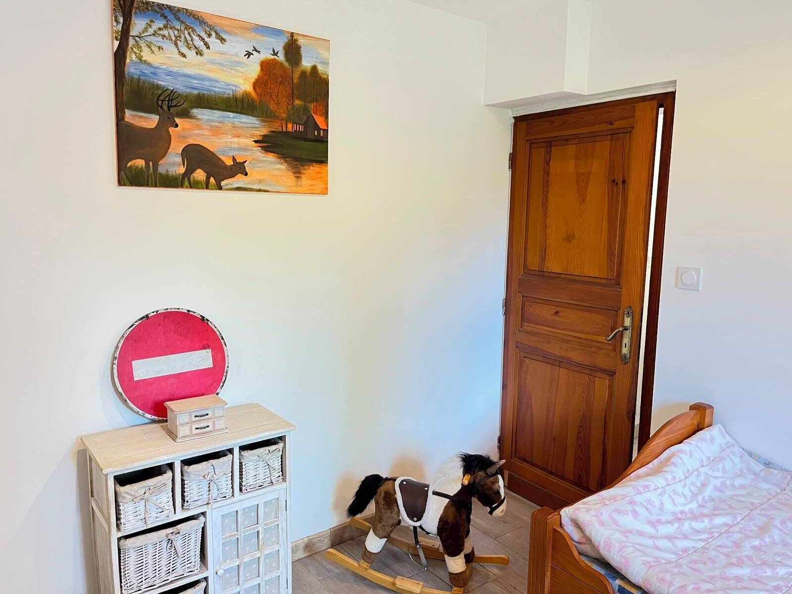 Una habitación con puerta de madera, una cama, un caballo balancín, un mueble de almacenaje con cestas y un cuadro de un paisaje con ciervos.