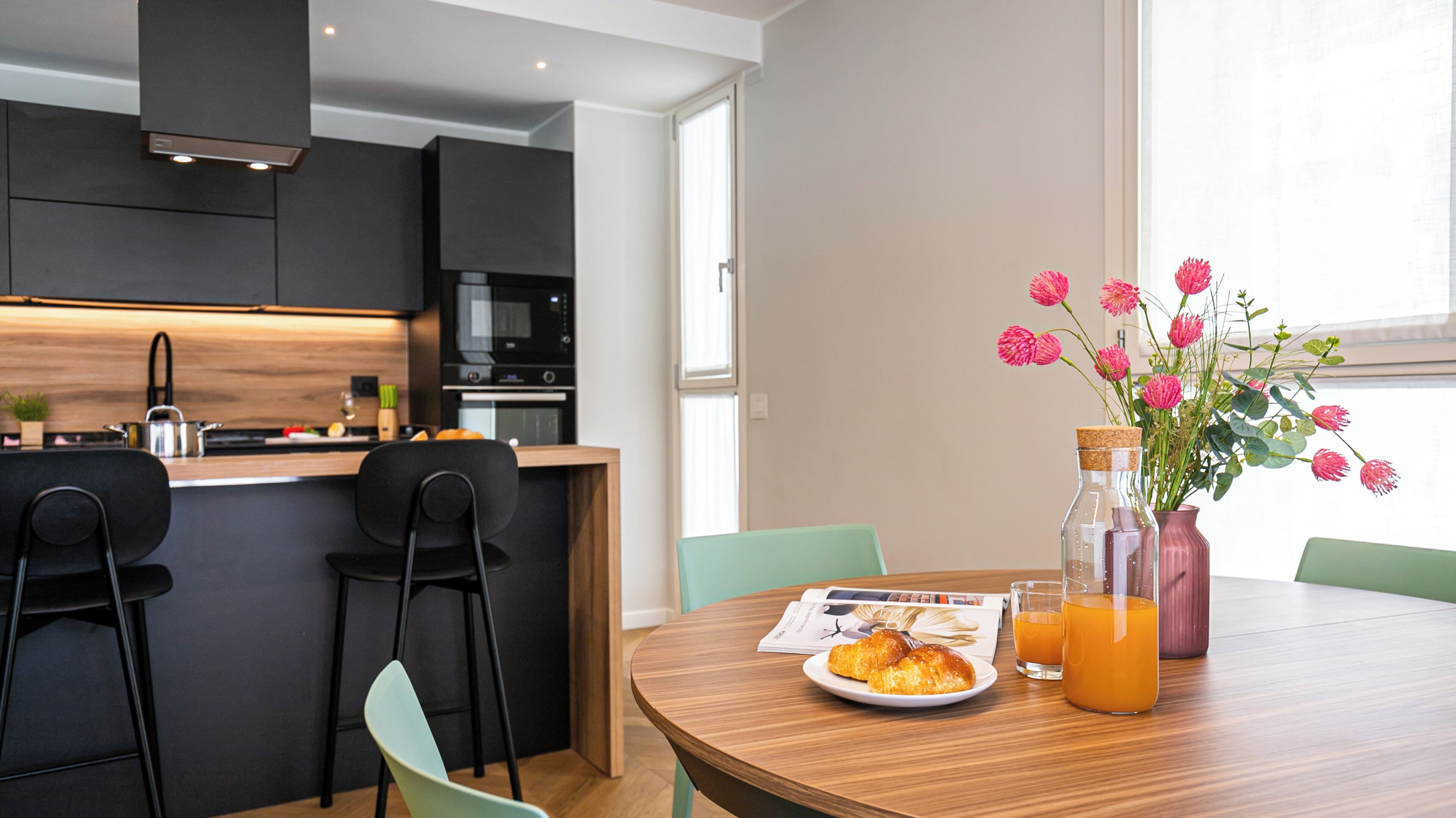 A modern kitchen with dark cabinetry, a wooden countertop, and a dining area with a round table, chairs, and a vase of flowers.