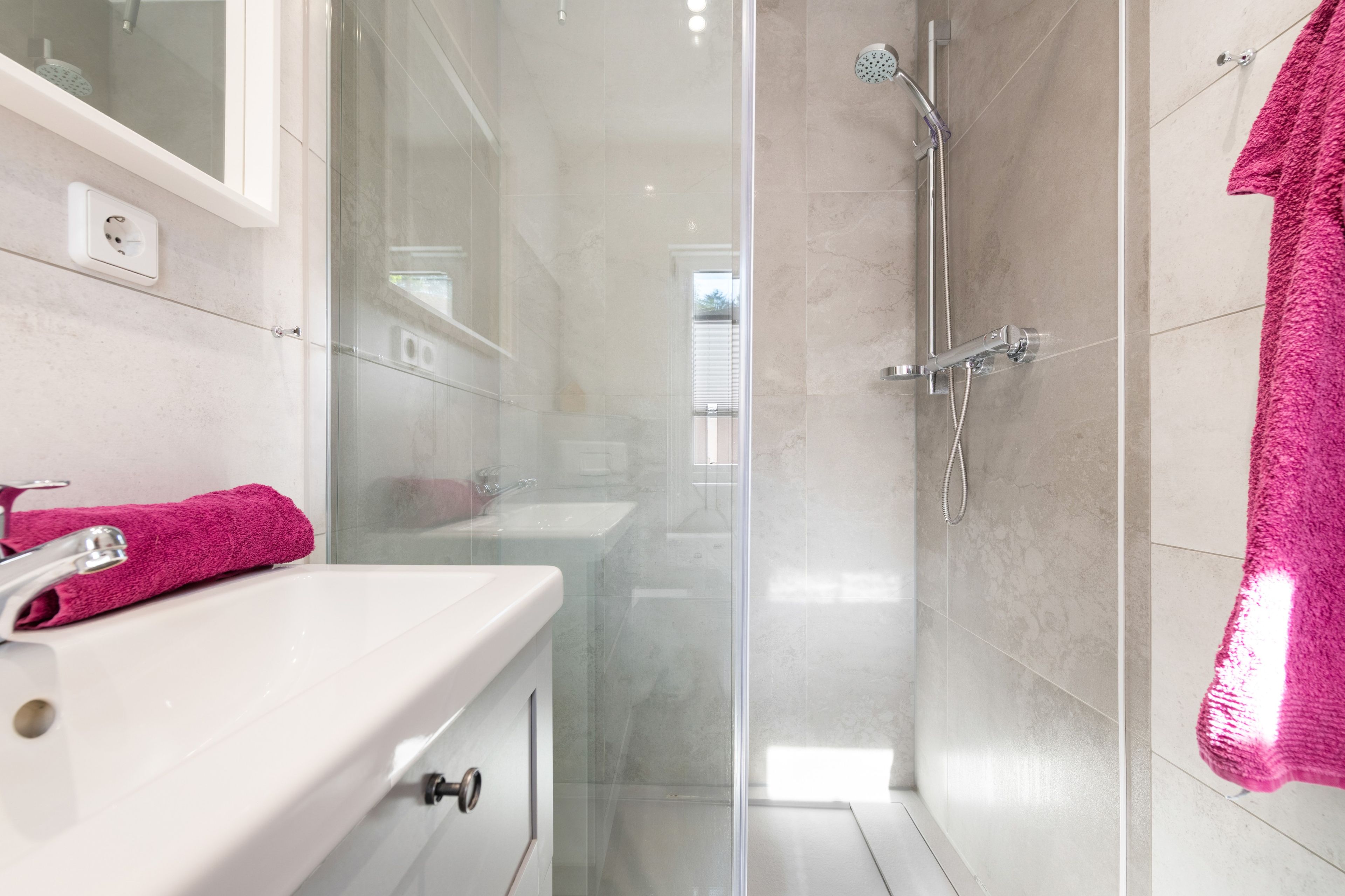 Ein modernes Badezimmer mit einer gläsernen Duschkabine, einem weißen Waschbecken und einem rosafarbenen Handtuch.