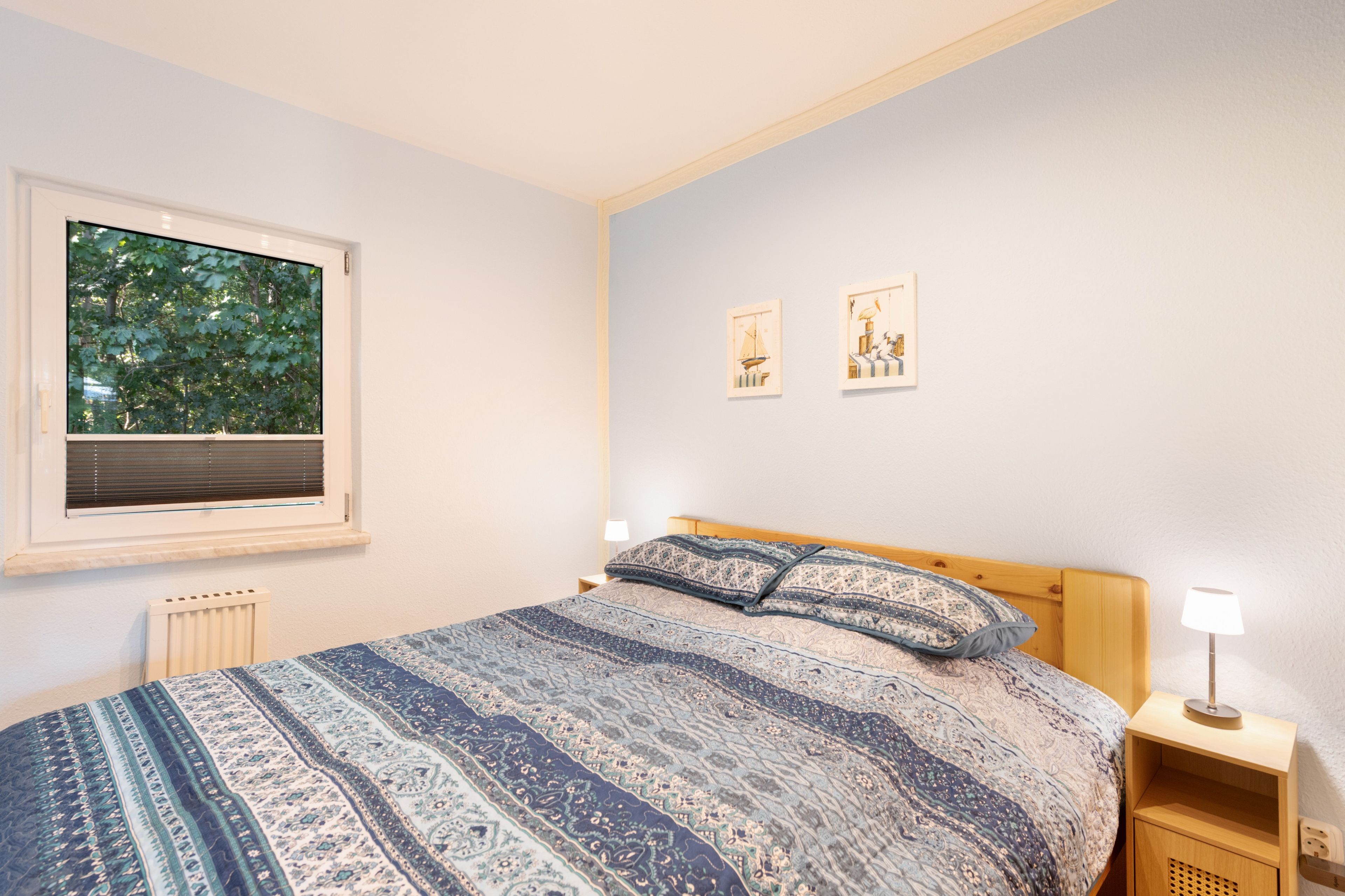 A bedroom with a large bed, a window with blinds, a radiator, two bedside tables with lamps, and two framed pictures on the wall.
