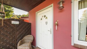 Una puerta blanca con un cartel de bienvenida, una silla de mimbre y un pequeño escalón que lleva a la entrada.