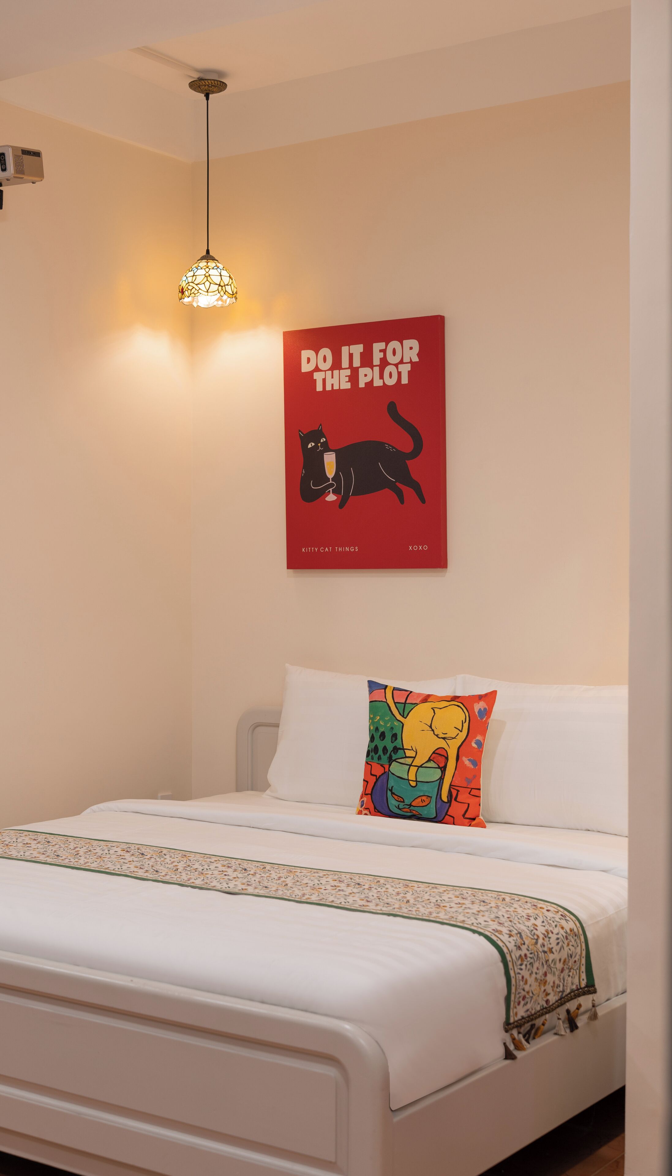 Ein Schlafzimmer mit einem Bett, einem farbenfrohen Kissen, einem roten Poster und einer Pendelleuchte.