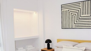 Um quarto moderno com cama, mesa de cabeceira, abajur e uma obra de arte geométrica na parede.