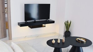 Uma sala de estar moderna com uma televisão de tela plana na parede, uma mesa de centro redonda preta com objetos decorativos e um sofá com almofadas estampadas.