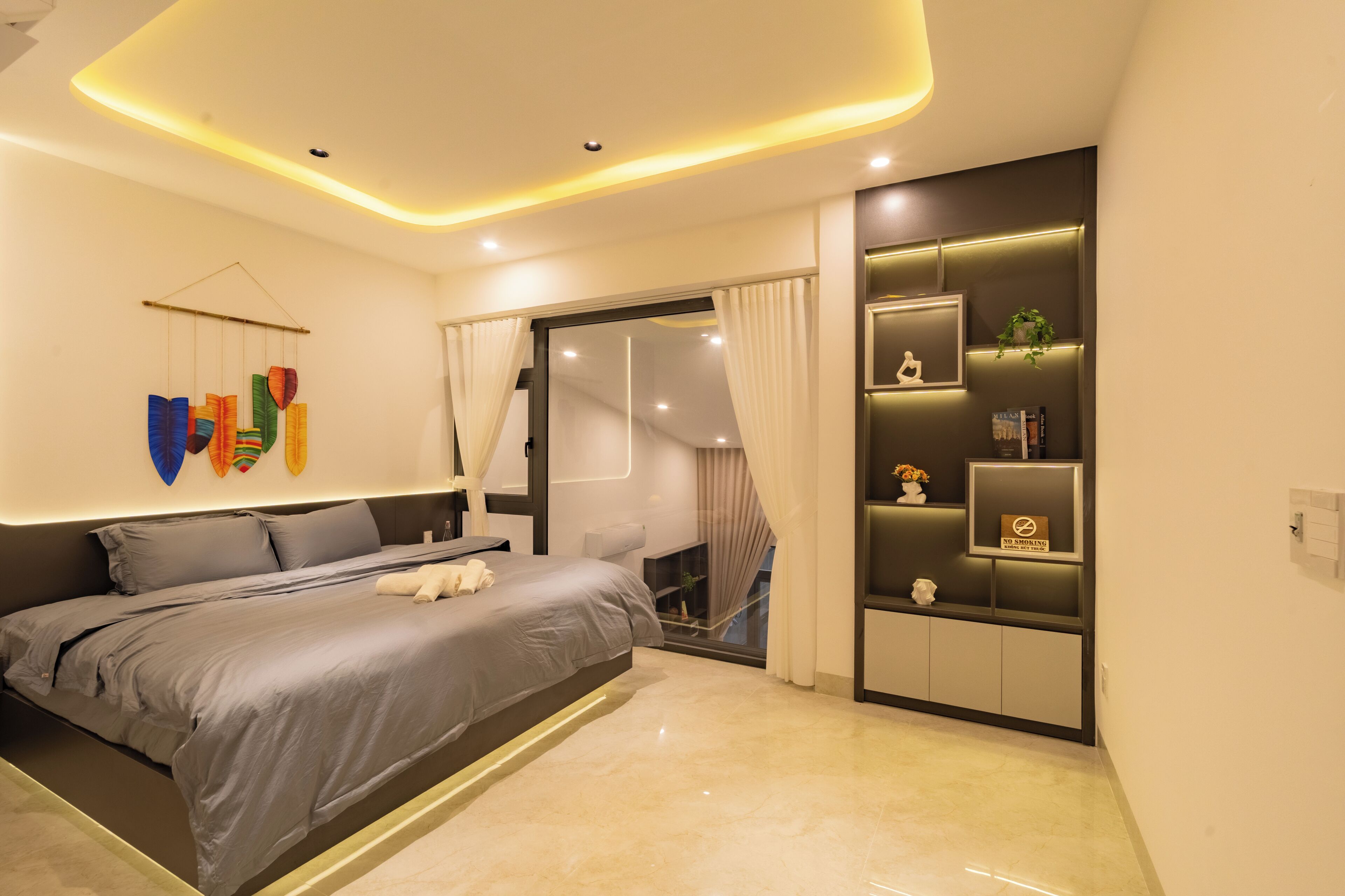 Een moderne slaapkamer met een bed, nachtkastjes, een wandplank met decoratieve objecten en uitzicht door een raam met gordijnen.