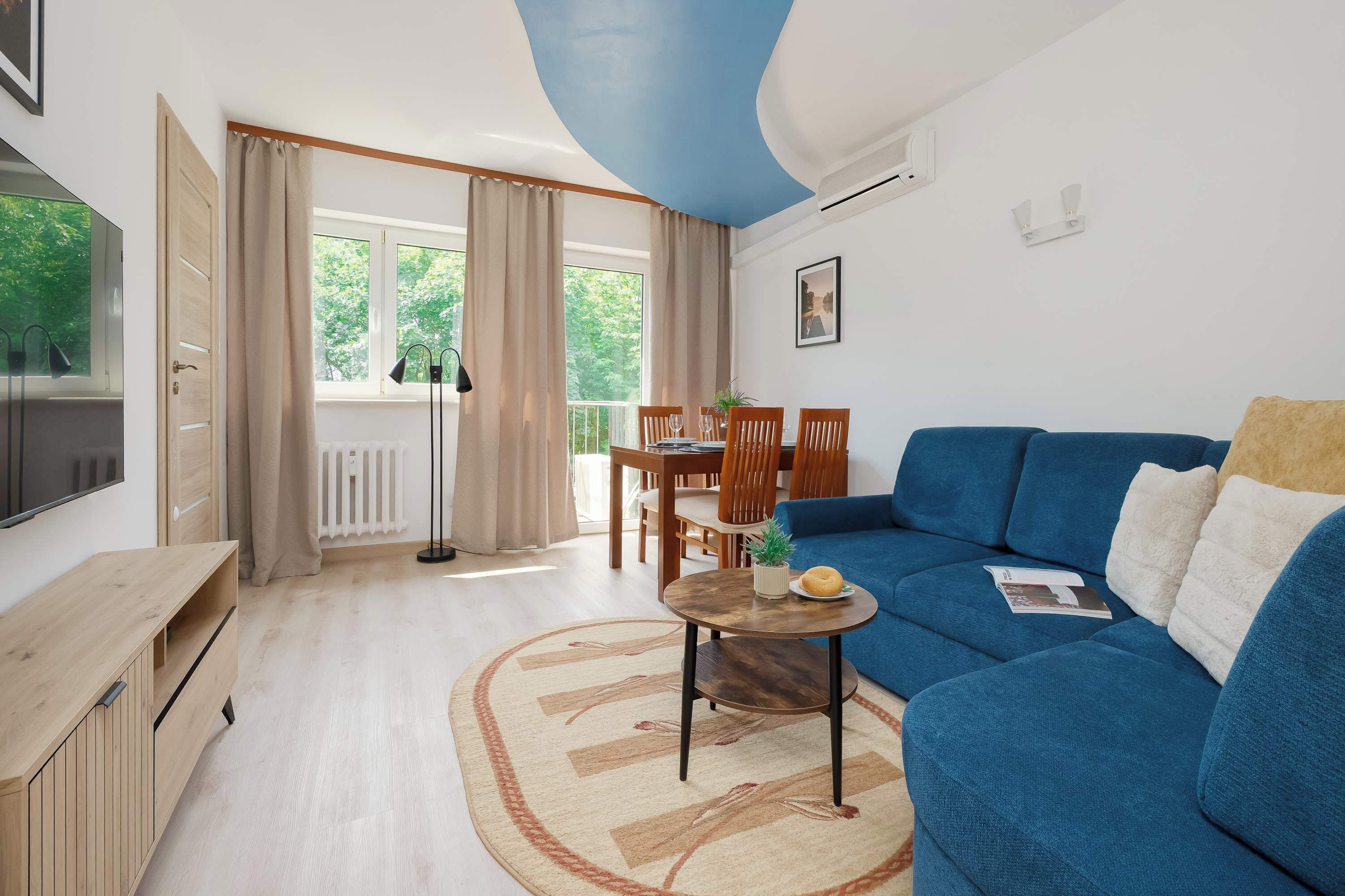 Uma sala de estar moderna com um sofá azul, uma mesa de jantar de madeira e uma TV à esquerda.