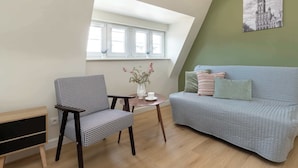 A room with a houndstooth patterned chair, a light blue sofa, a small round table with a white vase and a cup, and a large window with white frames. - Cosy Studio in the Downtown in Gdansk by Noclegi Renters (Gdańsk)