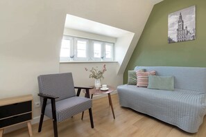 Ein Zimmer mit einem hundszahn-gemusterten Sessel, einem hellblauen Sofa, einem kleinen runden Tisch mit einer weißen Vase und einer Tasse sowie einem großen Fenster mit weißen Rahmen.