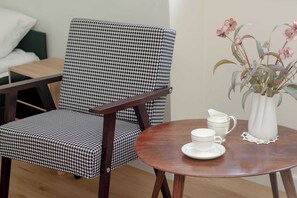 Ein Ohrensessel mit Hakensteinmuster, ein kleiner runder Holztisch mit weißer Karaffe, Tasse und Untertasse sowie eine Vase mit rosa Blumen.