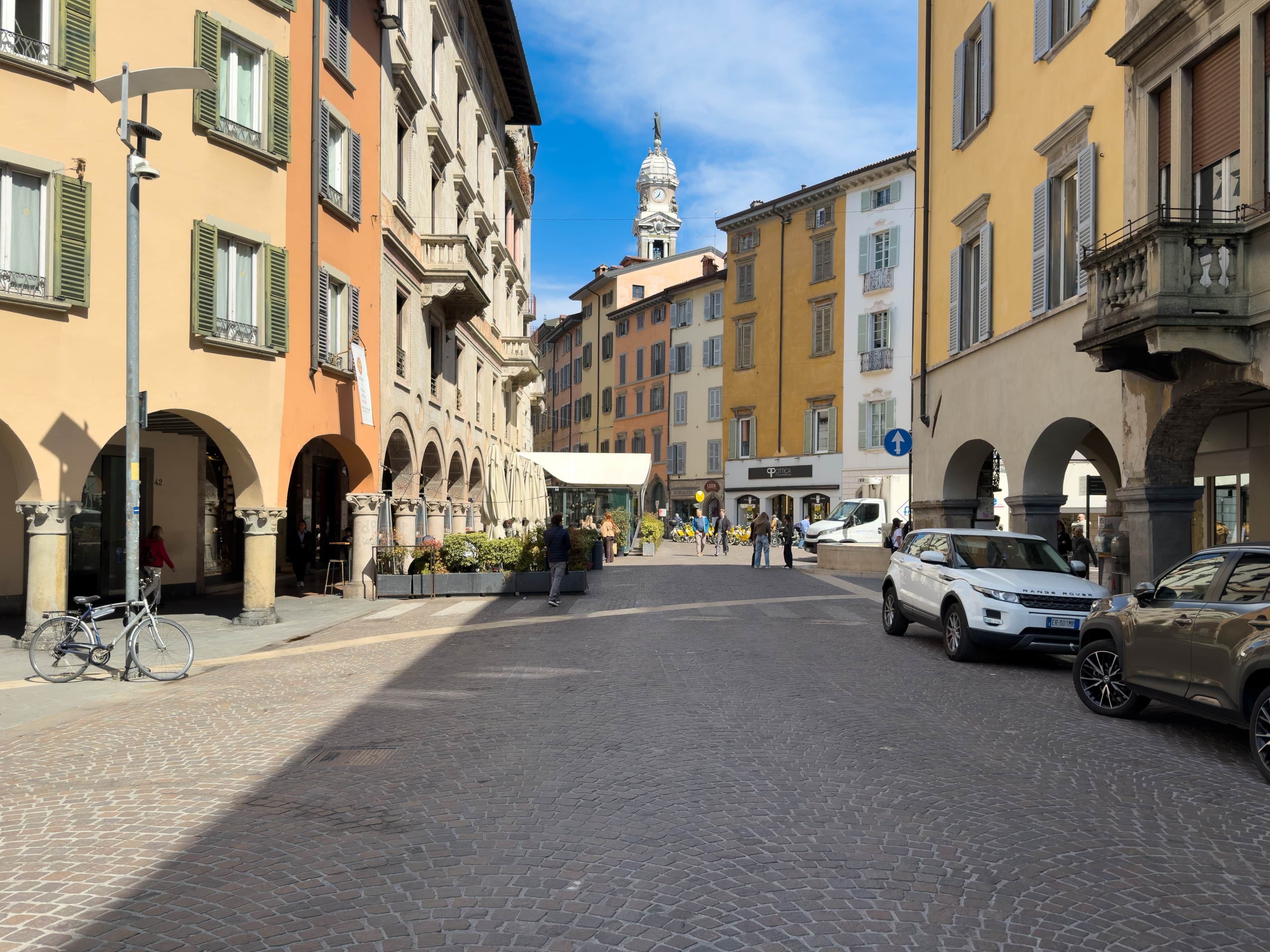 Una strada acciottolata fiancheggiata da edifici storici, una torre dell'orologio e auto parcheggiate.