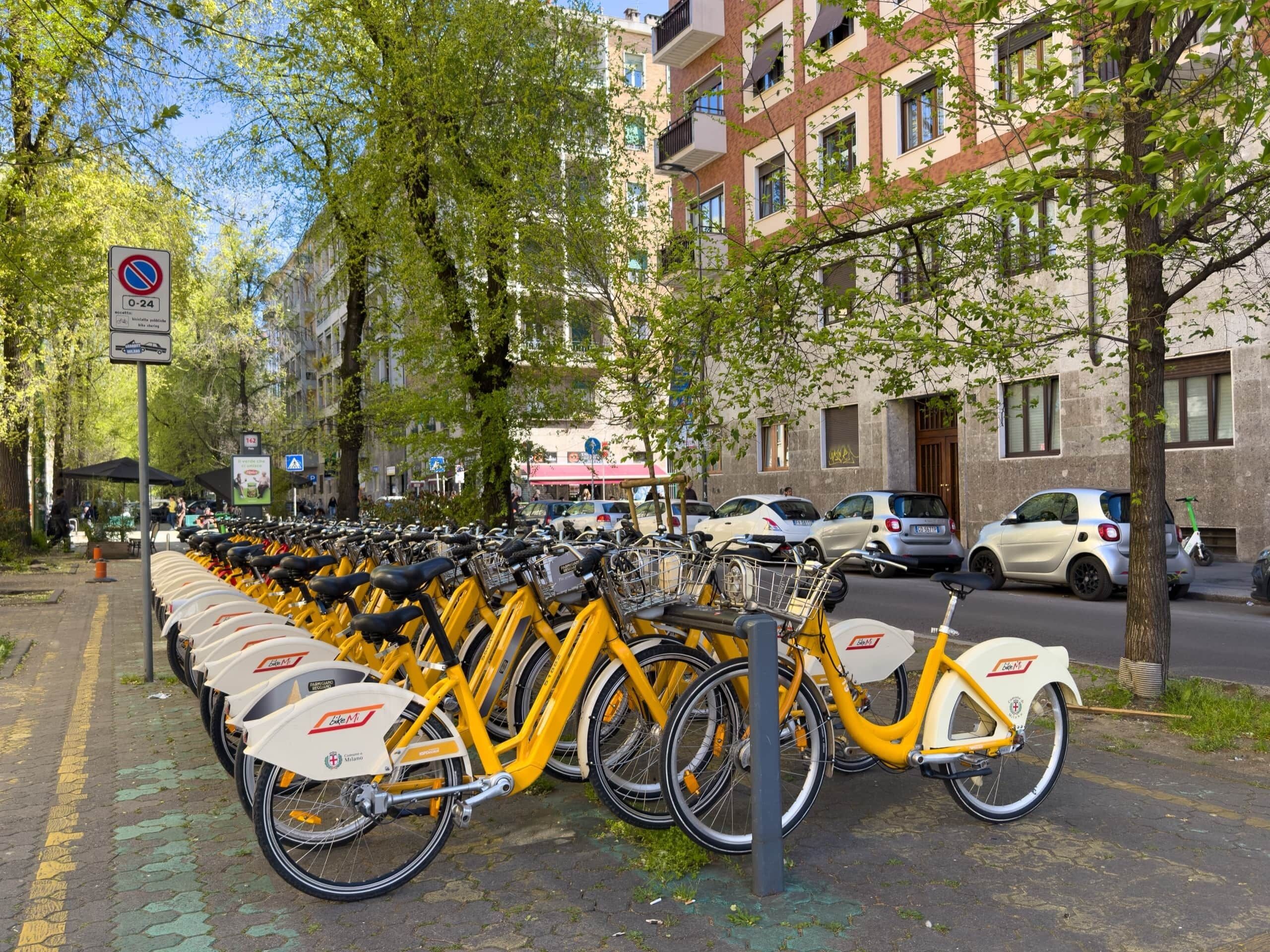 Una fila de bicicletas amarillas estacionadas en una acera, con un letrero de no estacionar, árboles y edificios al fondo.