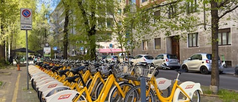 Una fila de bicicletas amarillas estacionadas en una acera, con un letrero de no estacionar, árboles y edificios al fondo.