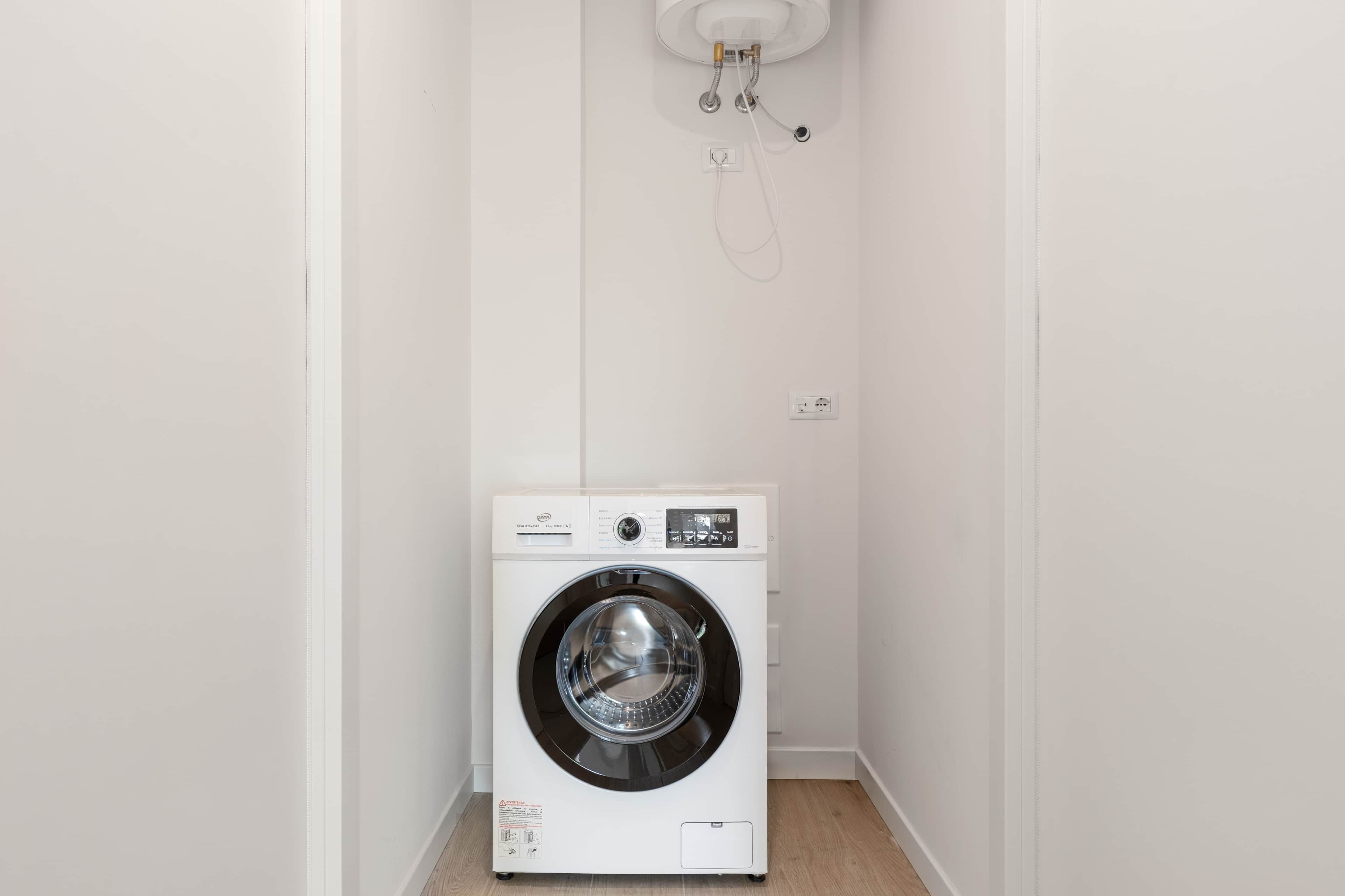 Une petite machine à laver blanche, fixée au mur dans un coin, avec un panneau de commande visible et une prise de courant.