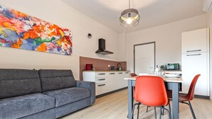 Ett modernt vardagsrum med en grå soffa, ett matbord med röda stolar, ett köksområde och en färgstark, abstrakt tavla på väggen.