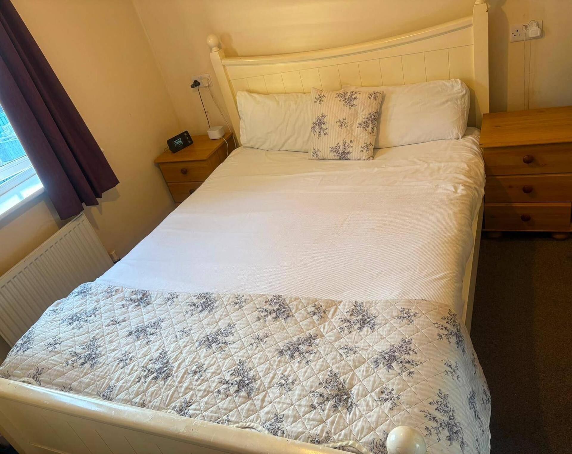 A single bed with a quilted bedspread and two pillows, a bedside table with a lamp, and a power outlet on the wall.