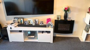 A living room with a flat-screen TV mounted on the wall, a white entertainment unit with various items, and a fireplace with a vase of flowers. - 3 Bedroom House Sleeps 5 -private Garden -parking (Leeds)