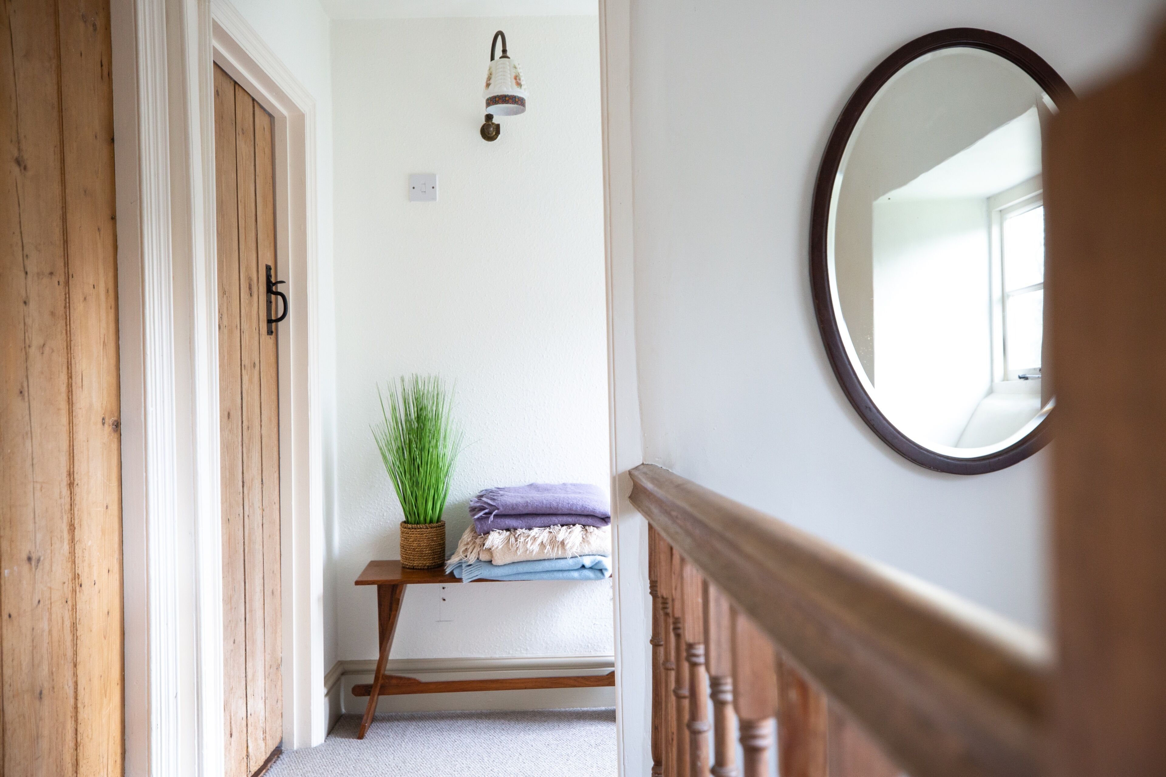 A hallway with a wooden door, a wall-mounted light fixture, a small table with a plant and folded towels, and a round mirror on the wall.