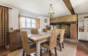 A dining area with a wooden table, wicker chairs, a chandelier, a fireplace, and a window with curtains.