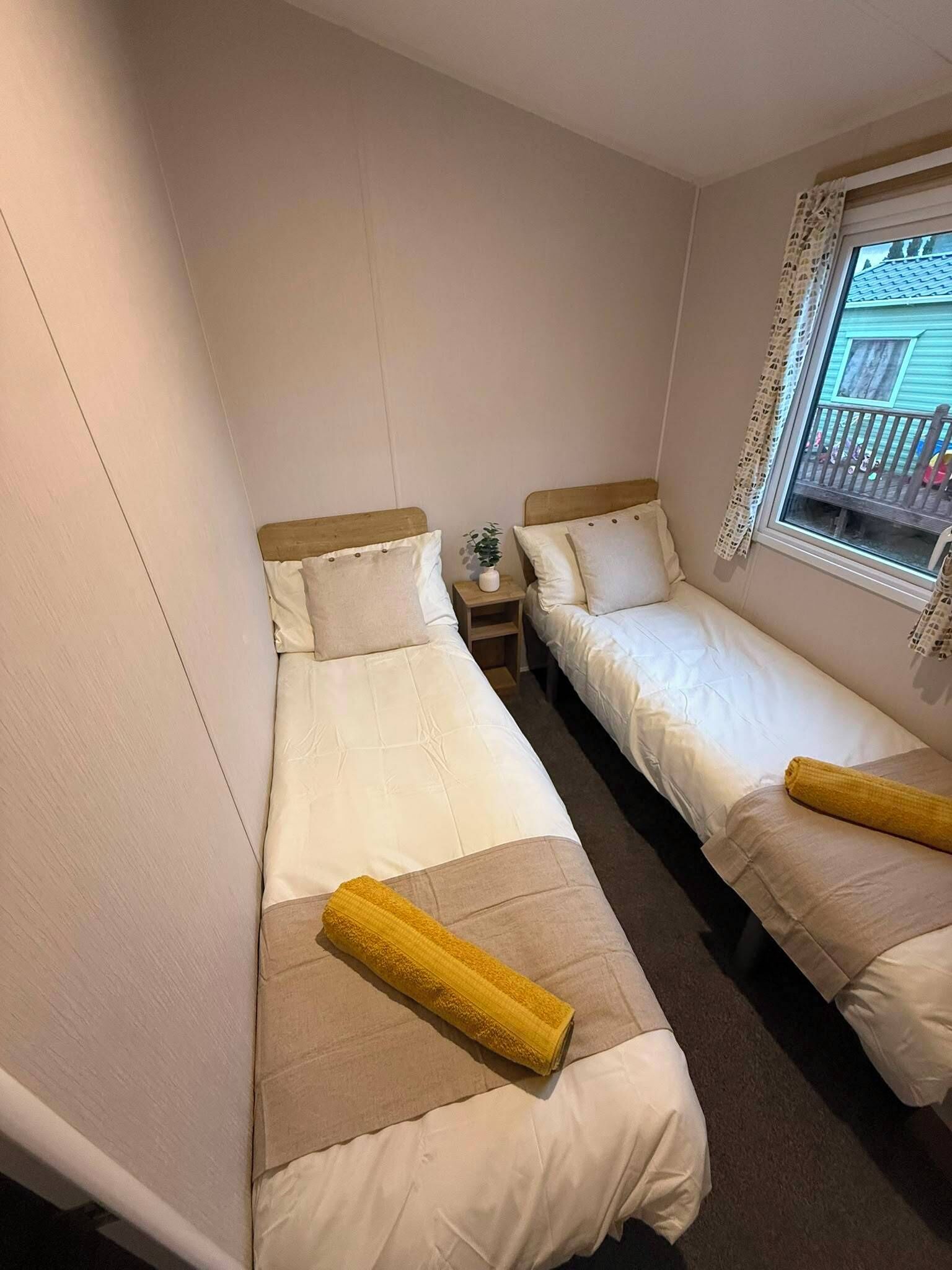 A small, compact room with two single beds, each with a pillow and a rolled-up towel. There is a small table with a plant between the beds.