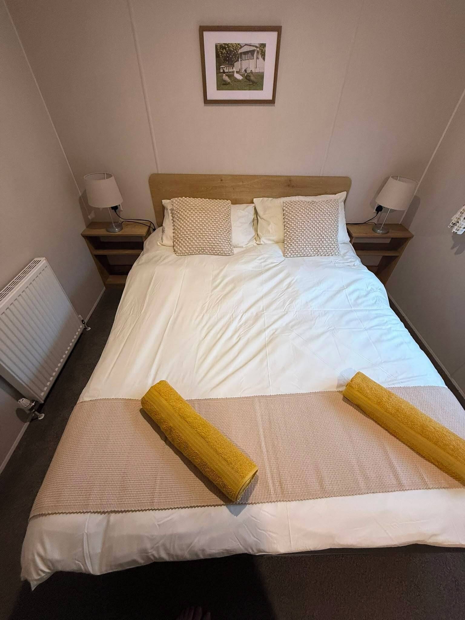 Una cama bien hecha con sábanas blancas y dos almohadas decorativas, un cabecero de madera y un cuadro en la pared.