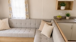 Uma sala de estar moderna com um sofá modular cinza, piso de madeira, área de cozinha com bancada de madeira e uma estante com plantas.