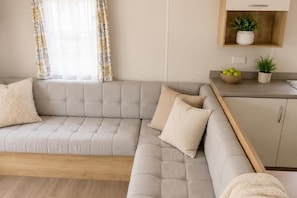 Ein modernes Wohnzimmer mit einem grauen Ecksofa, Holzboden, einer Küchenzeile mit Holzarbeitsplatte und einem Wandregal mit Pflanzen.