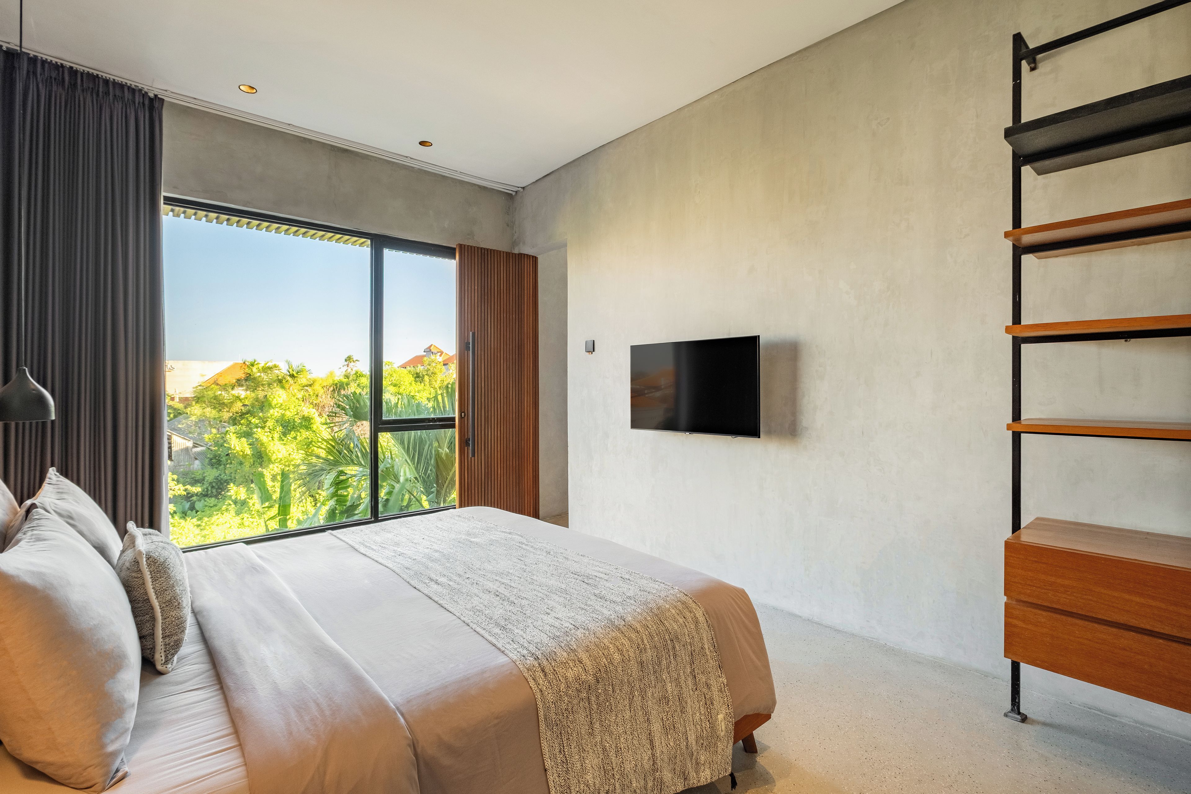 A bedroom with a large bed, a television mounted on the wall, and a view of greenery outside.