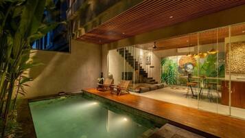 Un área de piscina interior moderna con terraza de madera, sillones de descanso y una escalera que lleva a una sala de estar con un mural.