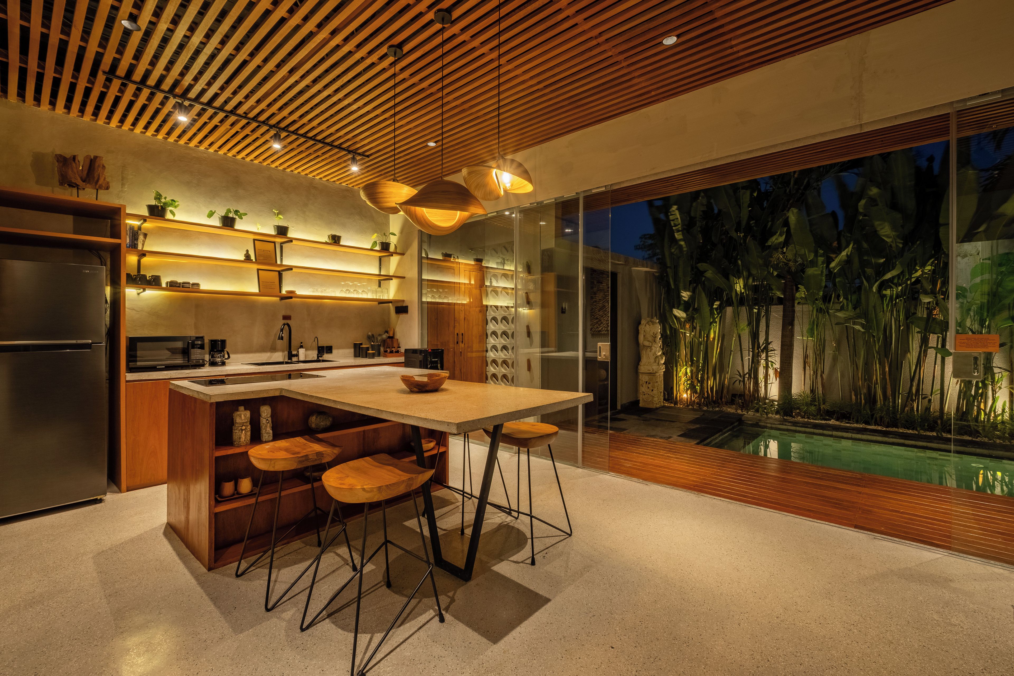 Una cocina moderna con una isla central, taburetes de bar y vista a una piscina por la noche.
