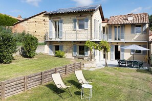 A two-story stone house with a fenced backyard, a small patio with a table and chairs, and a solar panel on the roof. - Cozy Court | Relaxation and a Jacuzzi in the Monferrato Hills (Grazzano Badoglio)