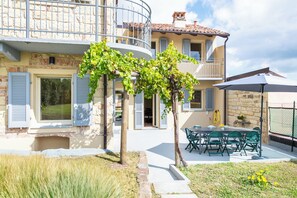 Ein zweigeschossiges Gebäude mit einem Balkon, einem Essbereich mit Gartenmöbeln und einem kleinen Garten mit einem Baum.