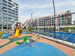 A playground with a slide and swings, surrounded by a fence and buildings in the background. - Bold Studio | Rooftop Pool & Spa | Oxford Arjan (Dubai)