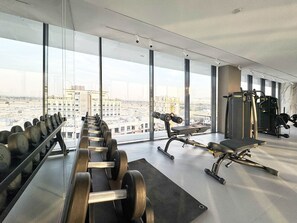 A gym with exercise equipment, large windows, and a view of the cityscape. - Bold Studio | Rooftop Pool & Spa | Oxford Arjan (Dubai)