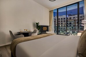 A hotel room with a large bed, a dining table with chairs, a small table with drinks, a plant, and a view of buildings through large windows. - Bold Studio | Rooftop Pool & Spa | Oxford Arjan (Dubai)
