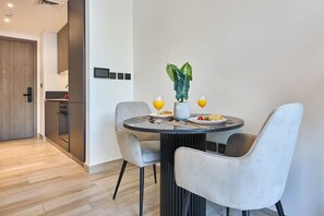 A modern dining area with a round table, two grey upholstered chairs, a potted plant, and a breakfast setting with orange juice glasses. - Bold Studio | Rooftop Pool & Spa | Oxford Arjan (Dubai)