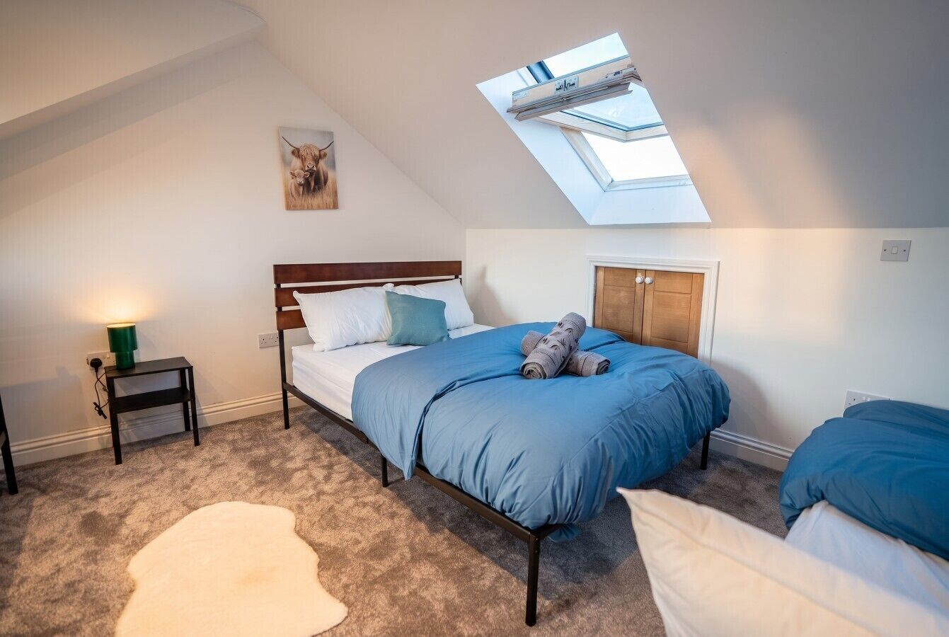 A bedroom with a skylight, a bed with blue bedding, a small bedside table, and a white furry rug.