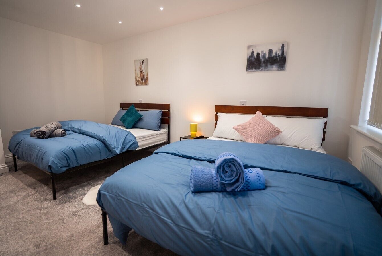 A hotel room with two beds, each with blue bedding and pillows, a nightstand with a lamp, and artwork on the walls.