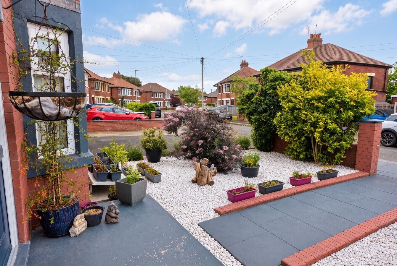 Een woonstraat met huizen, een oprit en een tuin met verschillende planten en een boom.