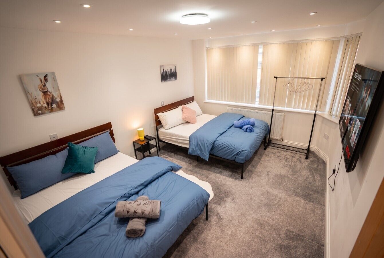 A hotel room with two beds, a window with blinds, a radiator, a small table with a lamp, and a wall-mounted TV.