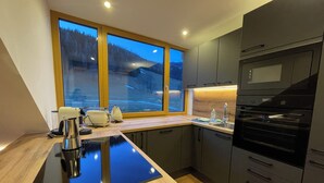 A modern kitchen with a large island, dark cabinetry, and a view of a snowy landscape through a large window. - Nockalm Cottage Apartment 5 Kaiserburg Superior (Sankt Oswald)