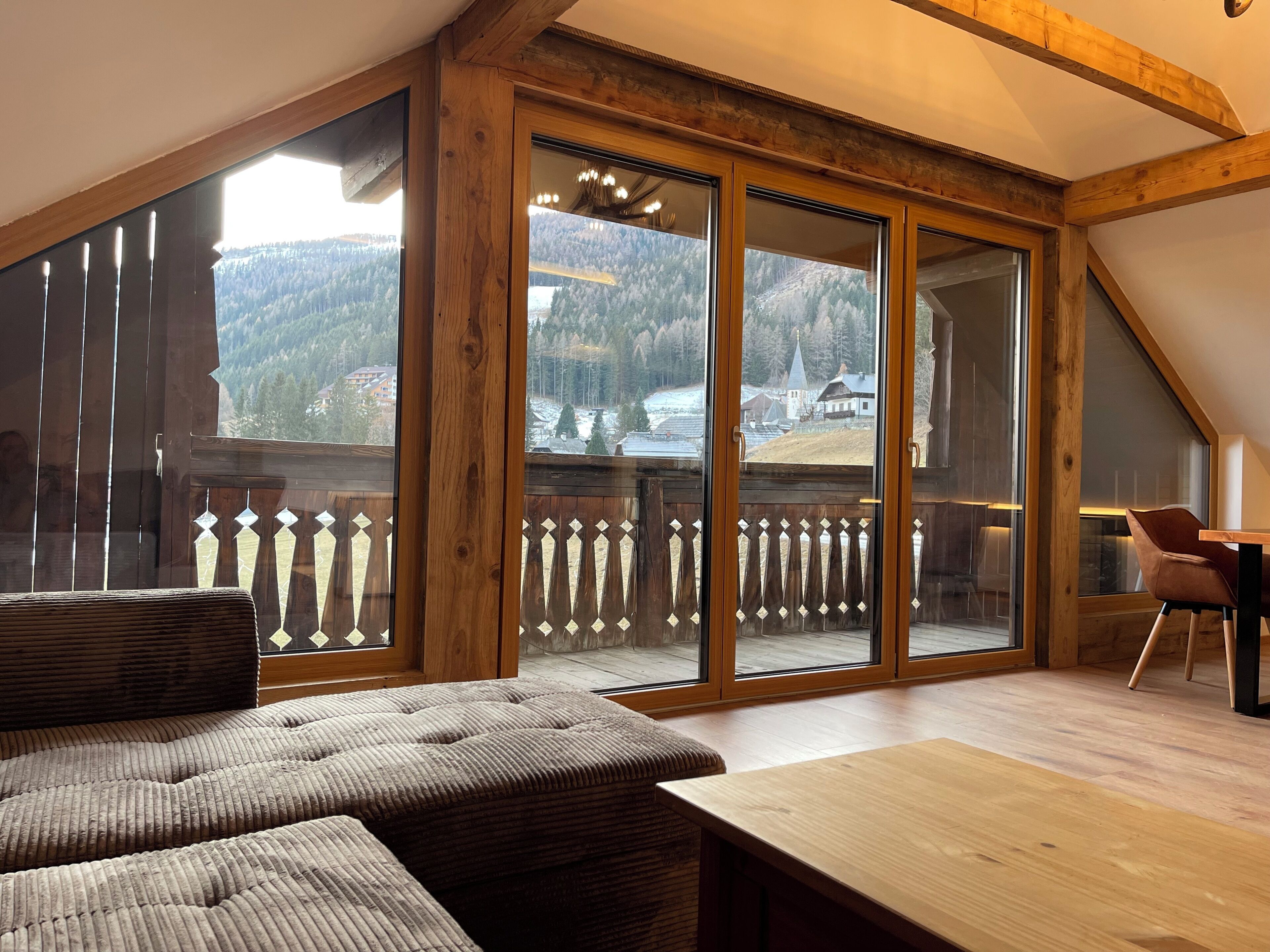 Una habitación con un amplio ventanal corredero que da a un balcón, una mesa de madera y un área de estar con cojines.
