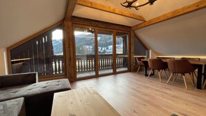 A spacious loft with a wooden ceiling, a dining area with chairs, a large window with a view of the mountains, and a chandelier. - Nockalm Cottage Apartment 5 Kaiserburg Superior (Sankt Oswald)