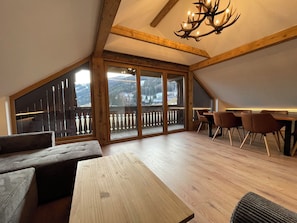 Ein geräumiges Loft mit Holzbalkendecke, einem Essbereich mit Stühlen, einem großen Fenster mit Bergblick und einer Chandelüster.