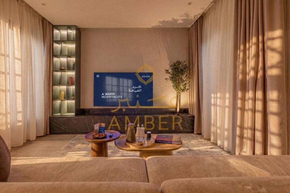A hotel lobby with a large screen displaying the hotel's name, a reception desk, a seating area with sofas and tables, and a plant.