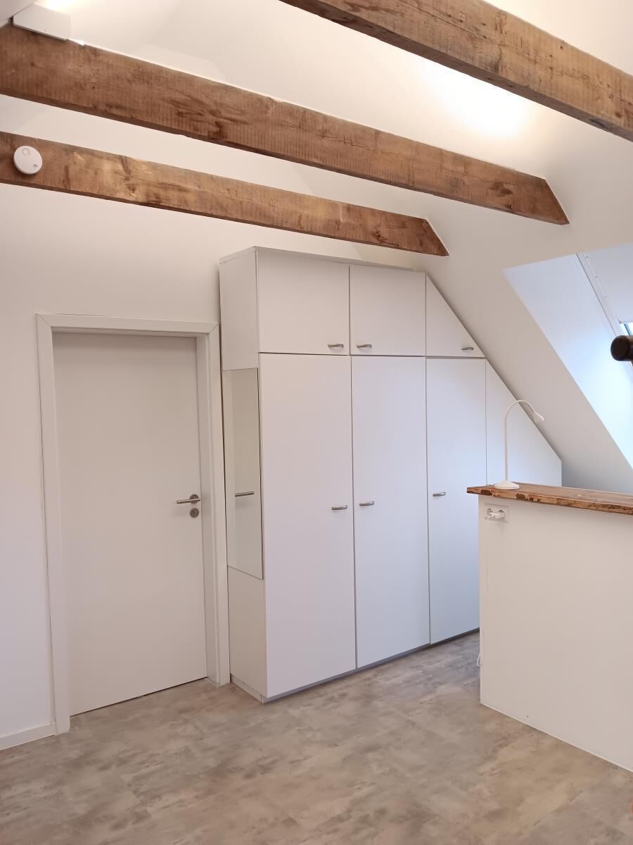 Ein Zimmer mit weißen Wänden, einem Holzbalken an der Decke, einem weißen Kleiderschrank und einer kleinen Holztheke.