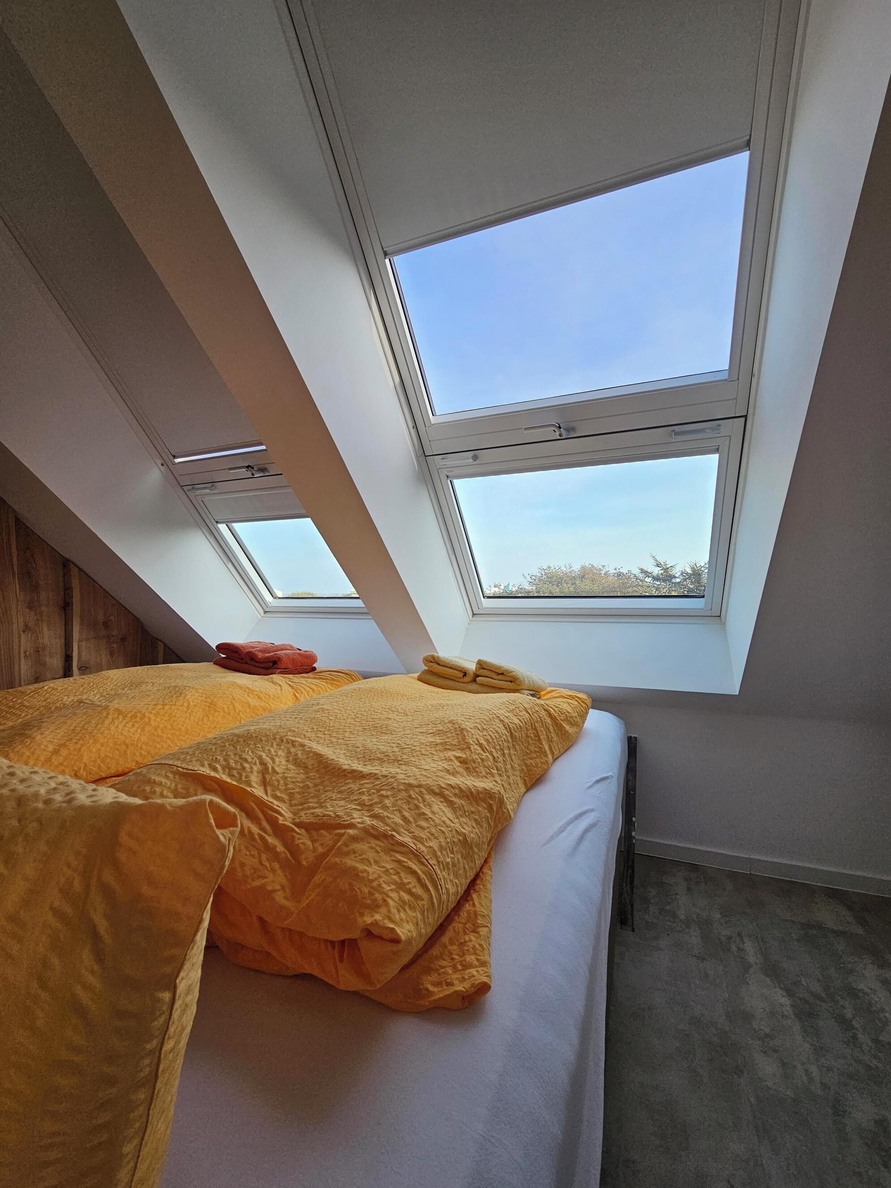 Une chambre avec un puits de lumière, un lit avec une literie jaune et une tête de lit en bois.