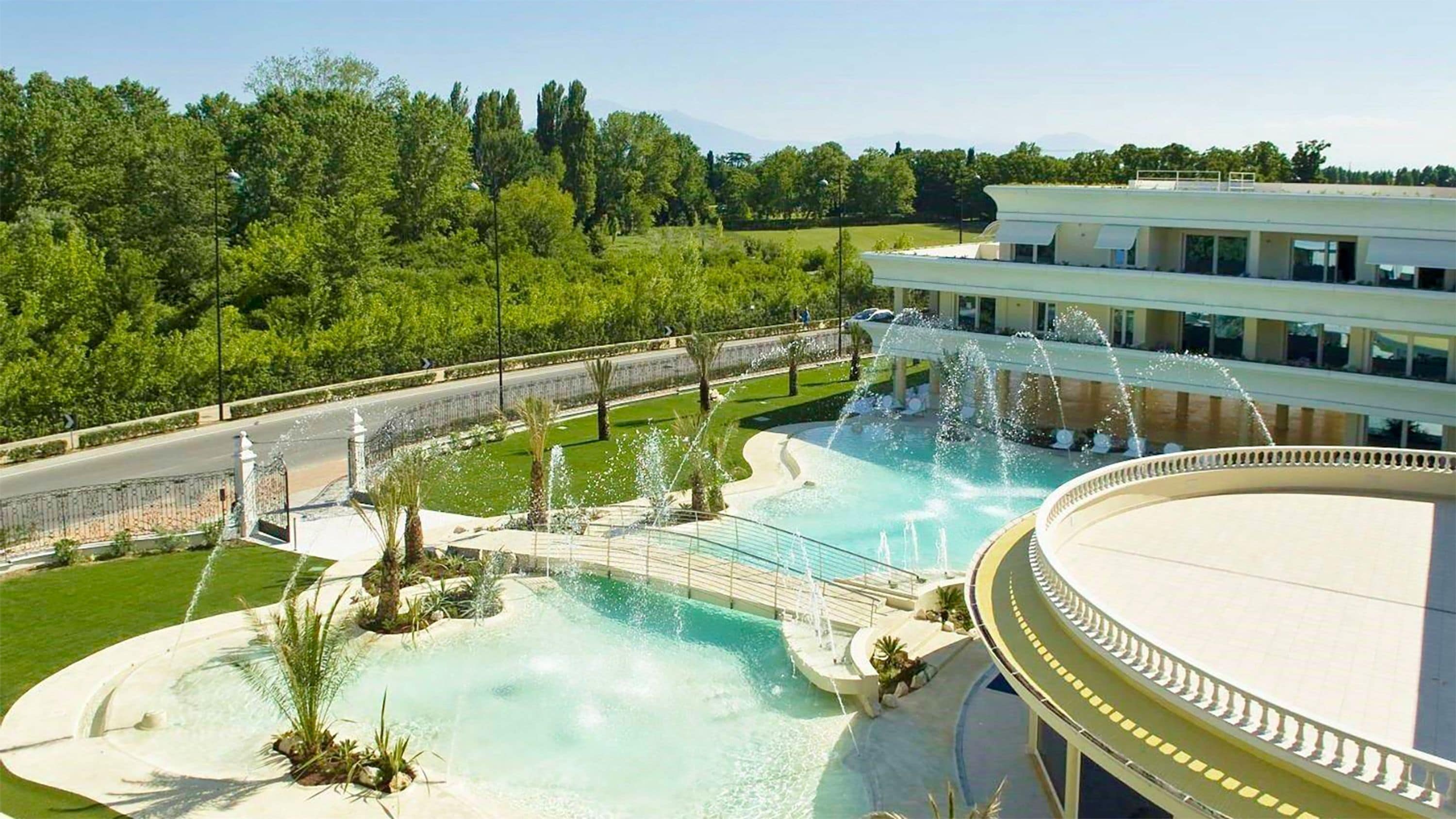 Une grande piscine extérieure agrémentée de plusieurs jeux d’eau, entourée de pelouses bien entretenues et d’arbres.