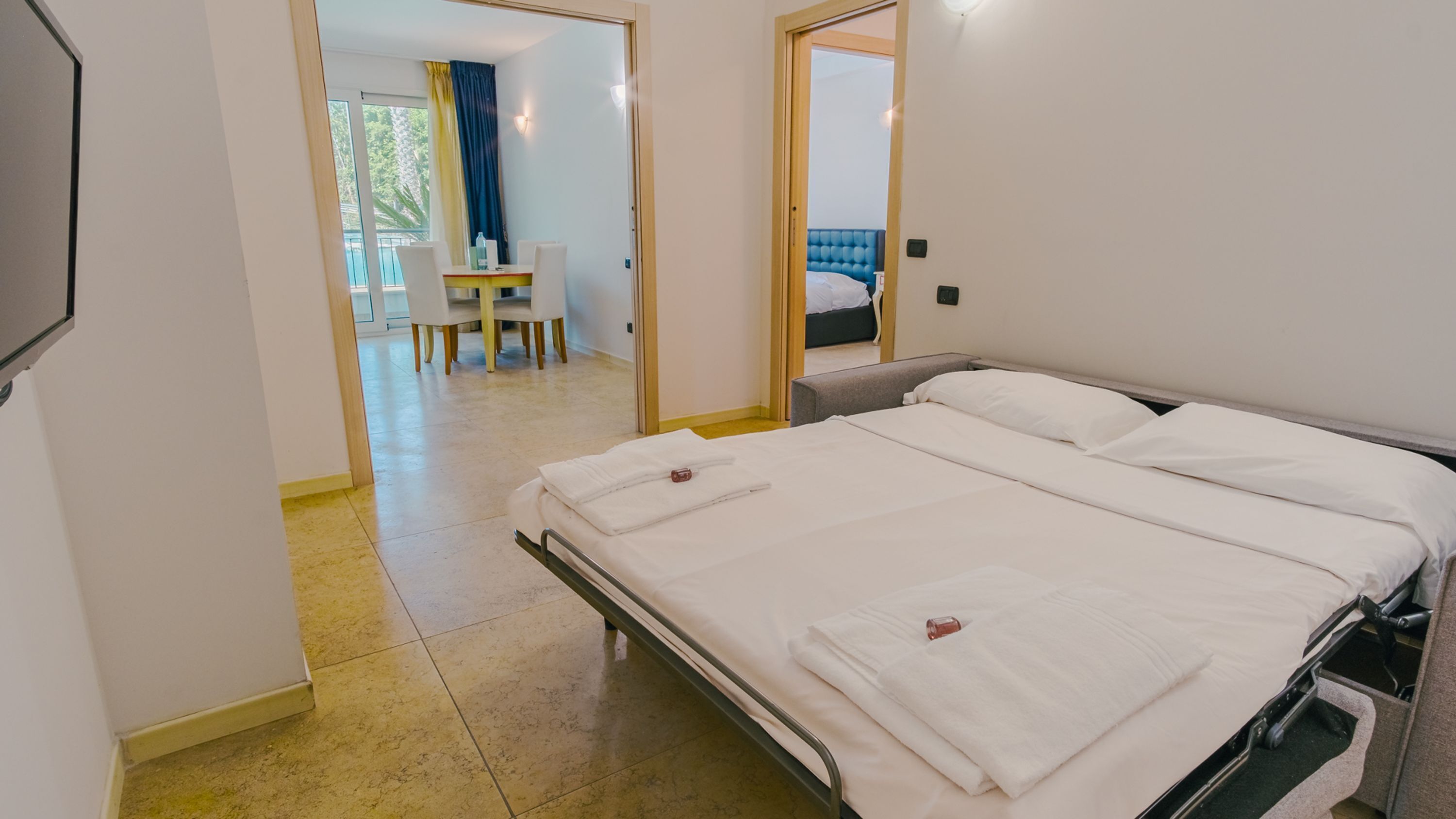 A hotel room with a double bed, a small table with chairs, and a view of a balcony.