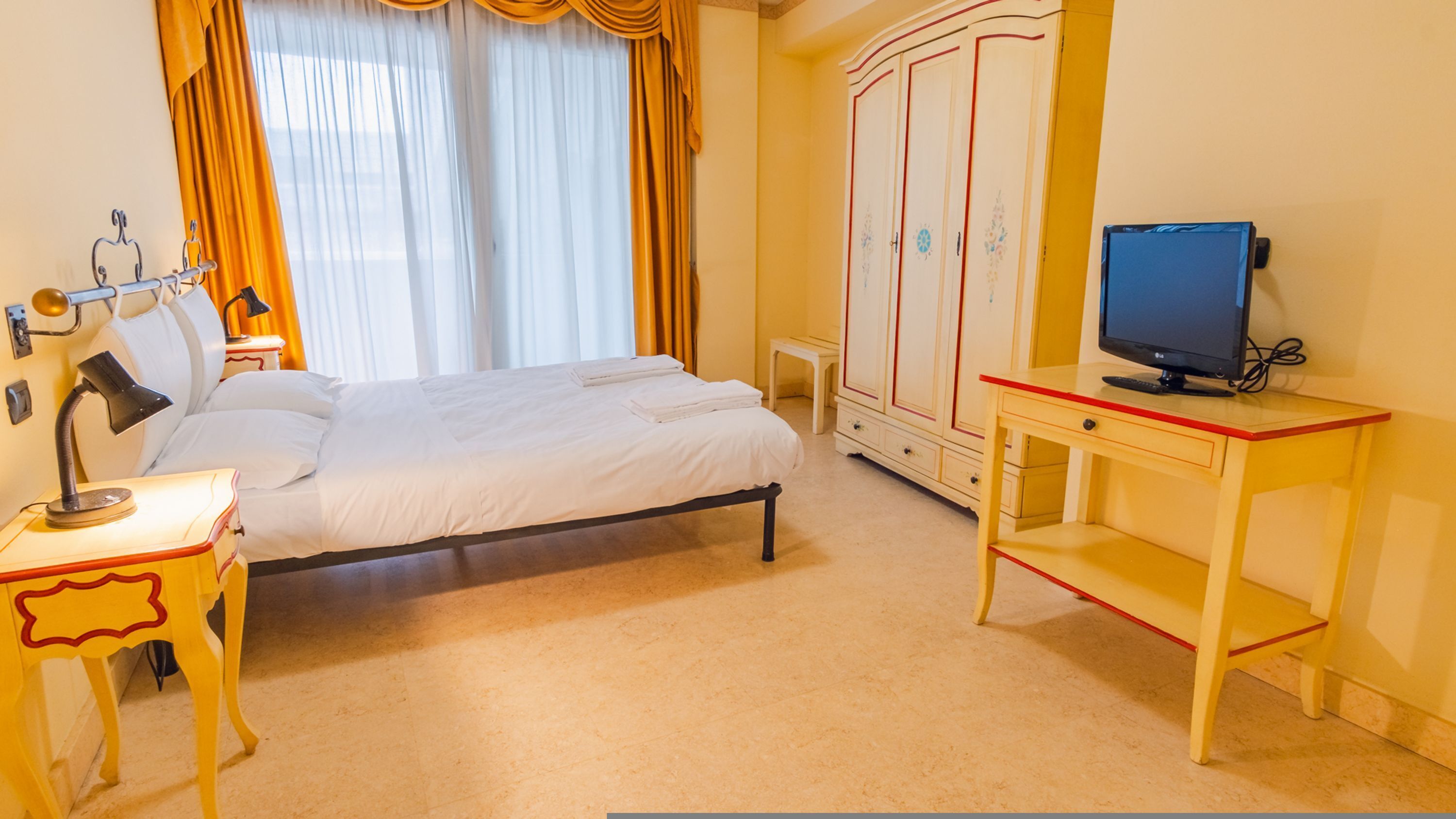 A hotel room with a bed, a desk with a computer, a wardrobe, and a window with curtains.
